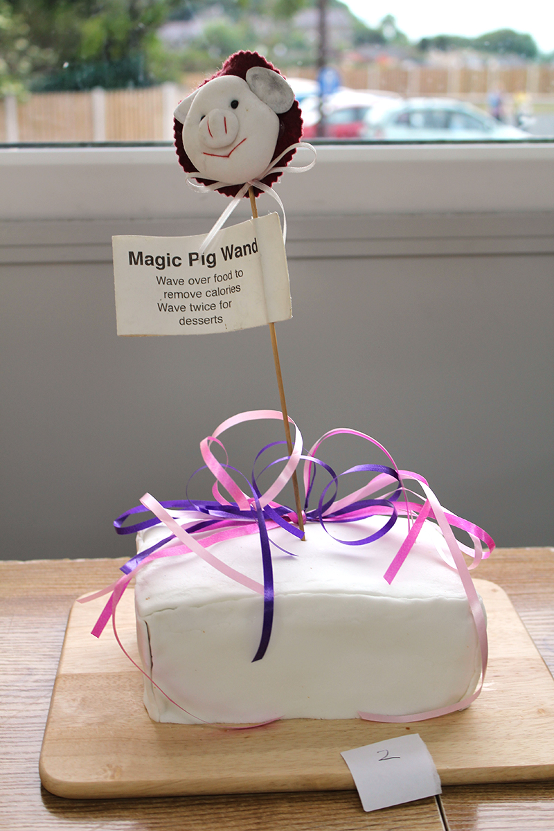Iced fruit cake with a pig wand struck in it, sign on wand promising that wand can remove calories when waved over the cake