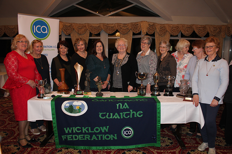 Prize Winners stand with their trophies and Cups on a table infront of them and the Wicklow Federation Banner draped over the table. Madge Kenny is in the centre of the group